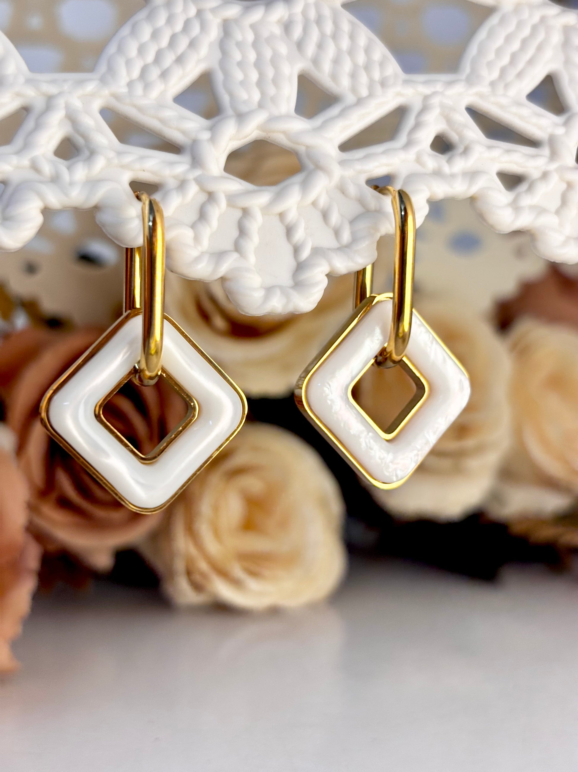 Gold and white square-shaped earrings on a decorative background with flowers.