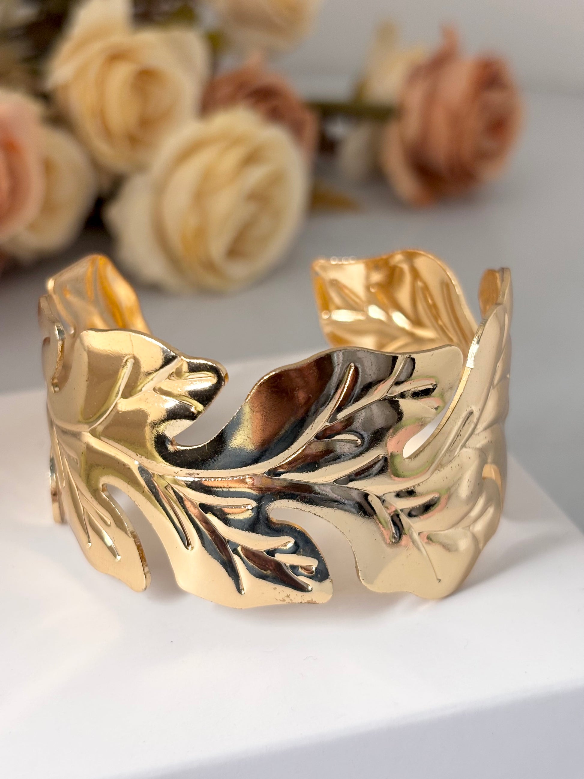 Gold leaf-patterned cuff bracelet on a white surface with flowers in the background