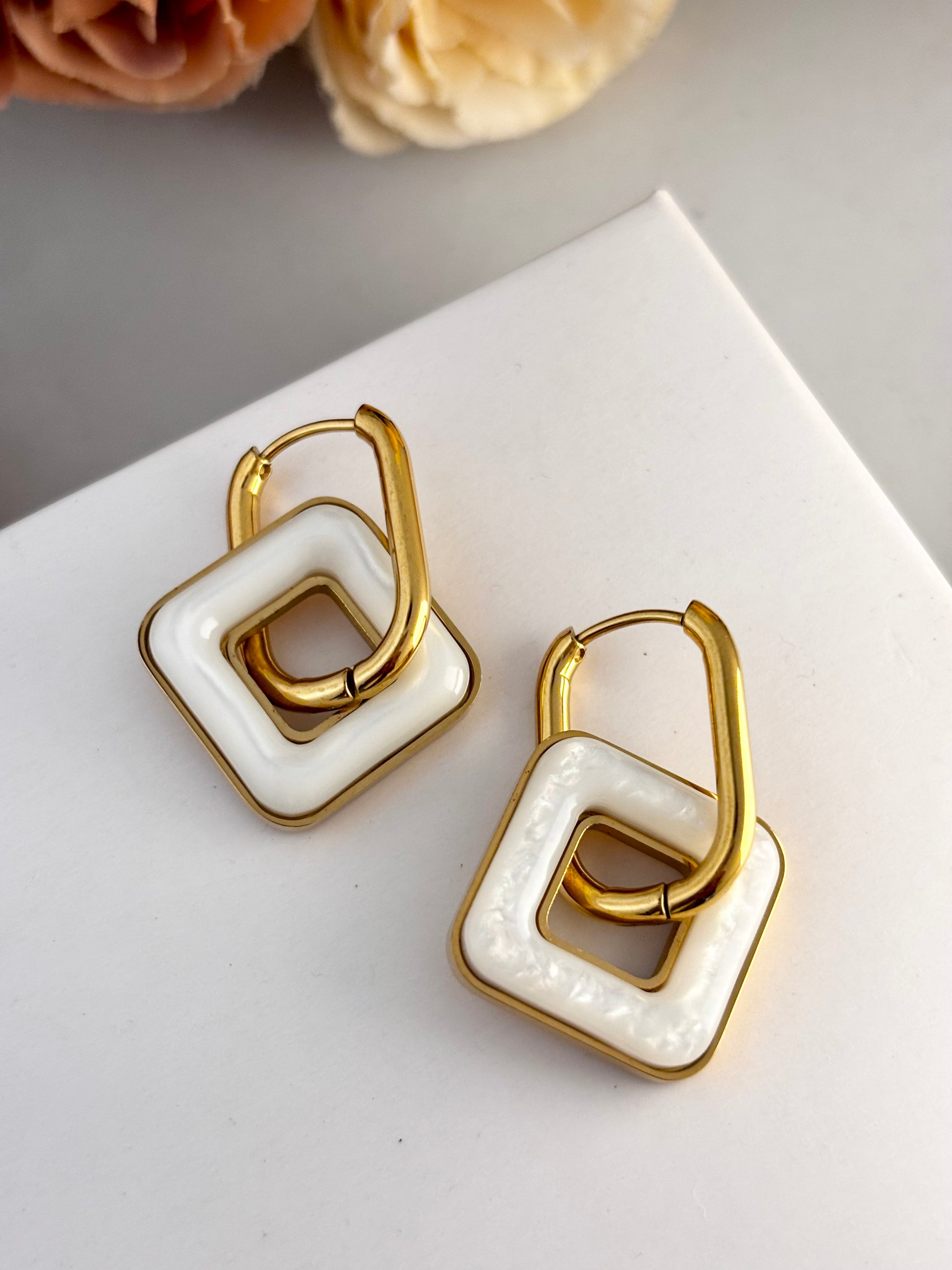 Gold and white square hoop earrings on a light surface with flowers in the background