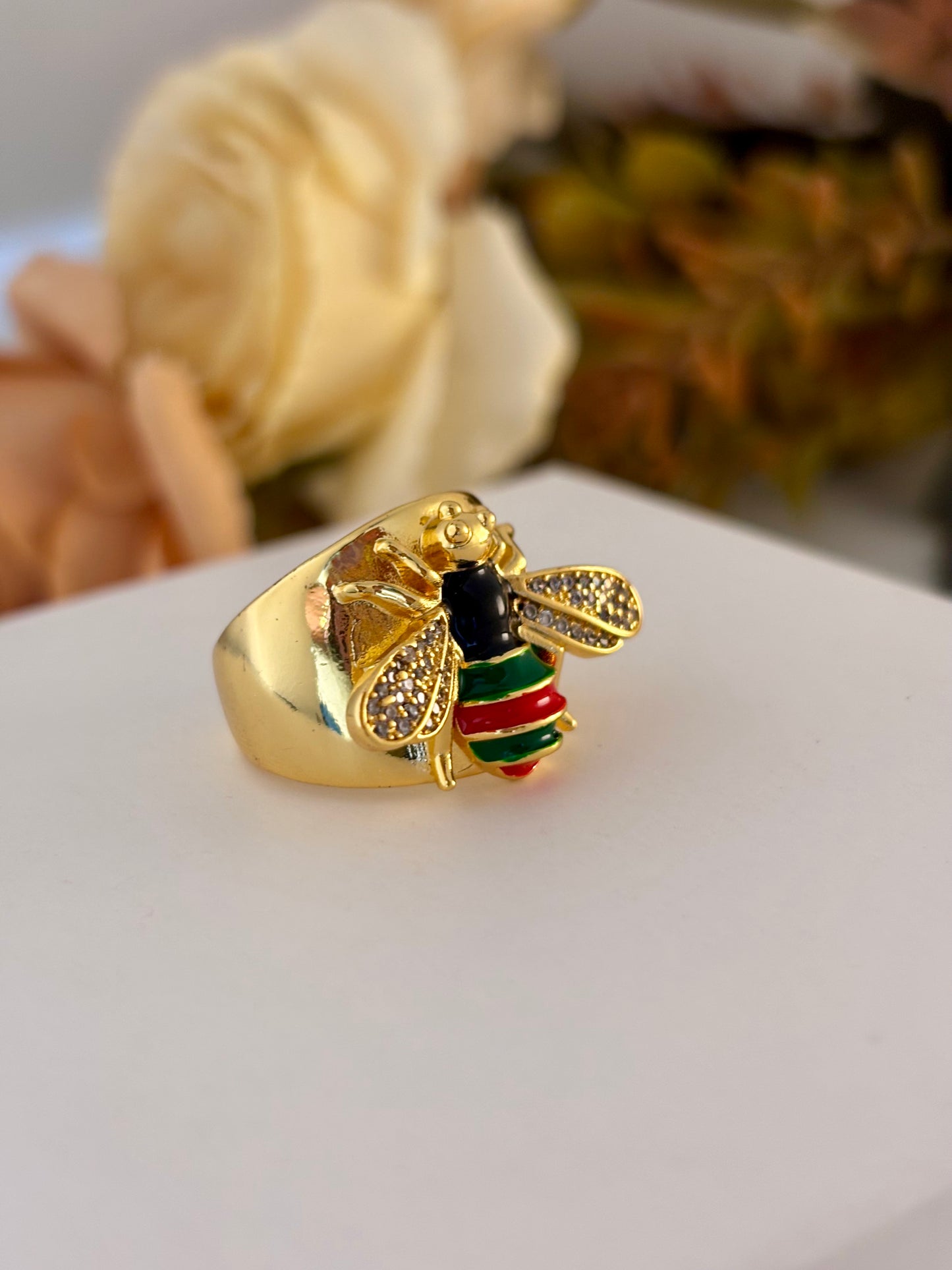 Gold bee-shaped ring with green and red accents on a white surface
