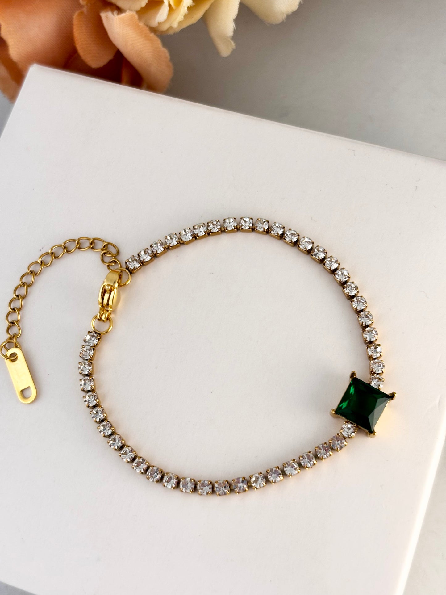 Gold bracelet with a green gemstone on a white surface with flowers in the background