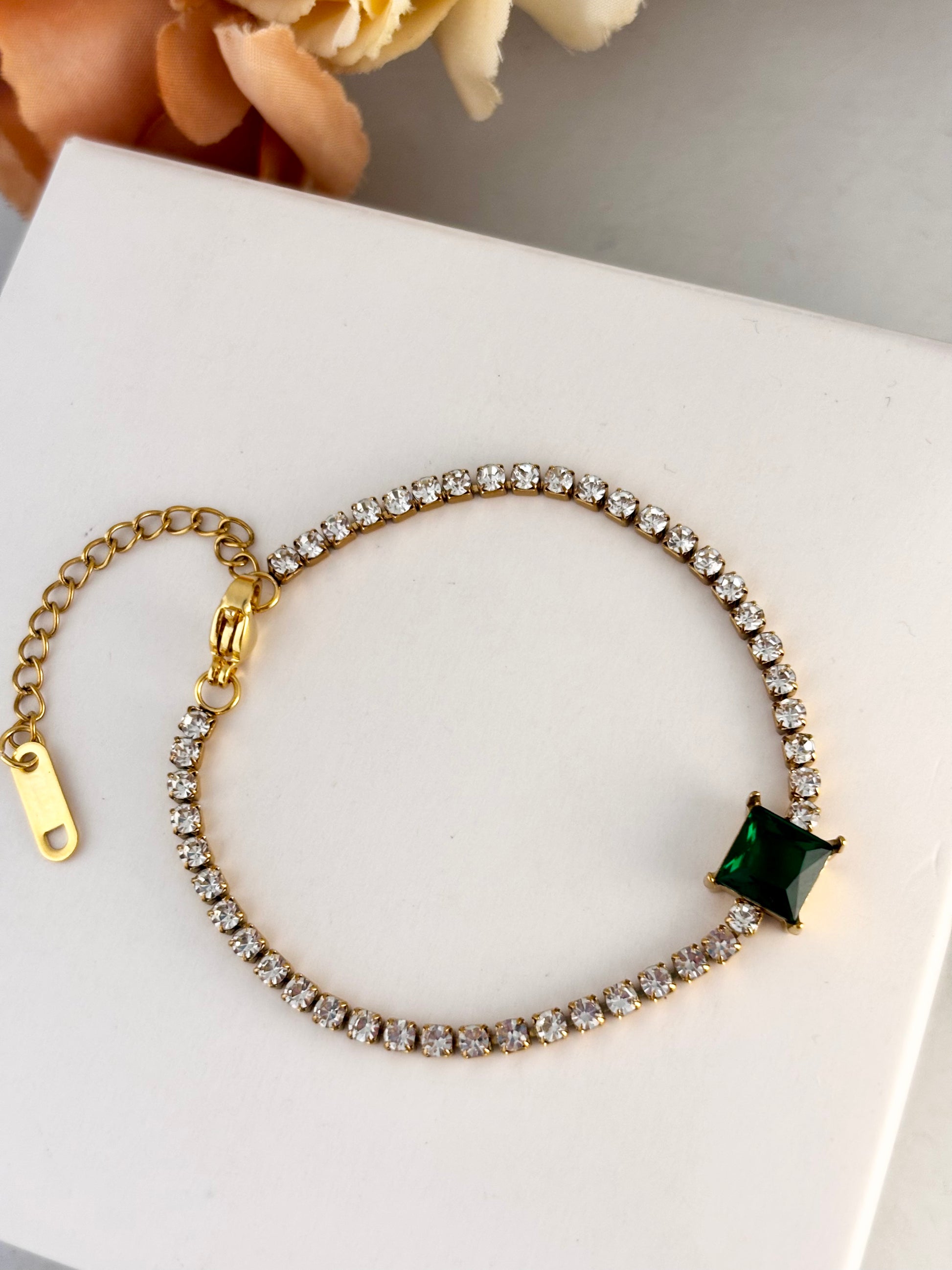 Gold bracelet with a green gemstone on a white surface with flowers in the background