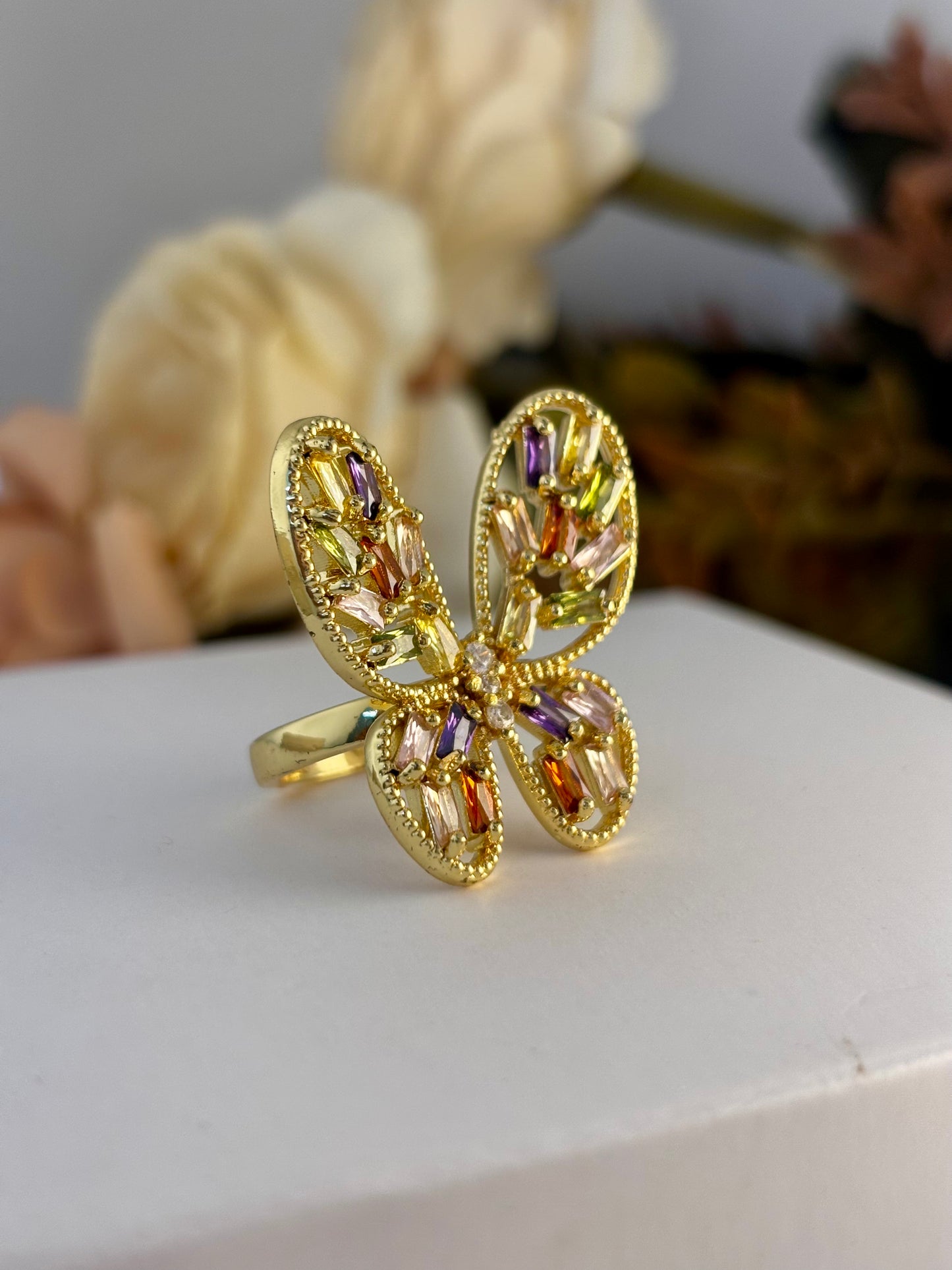 Gold butterfly ring with colorful gemstones on a white surface with flowers in the background