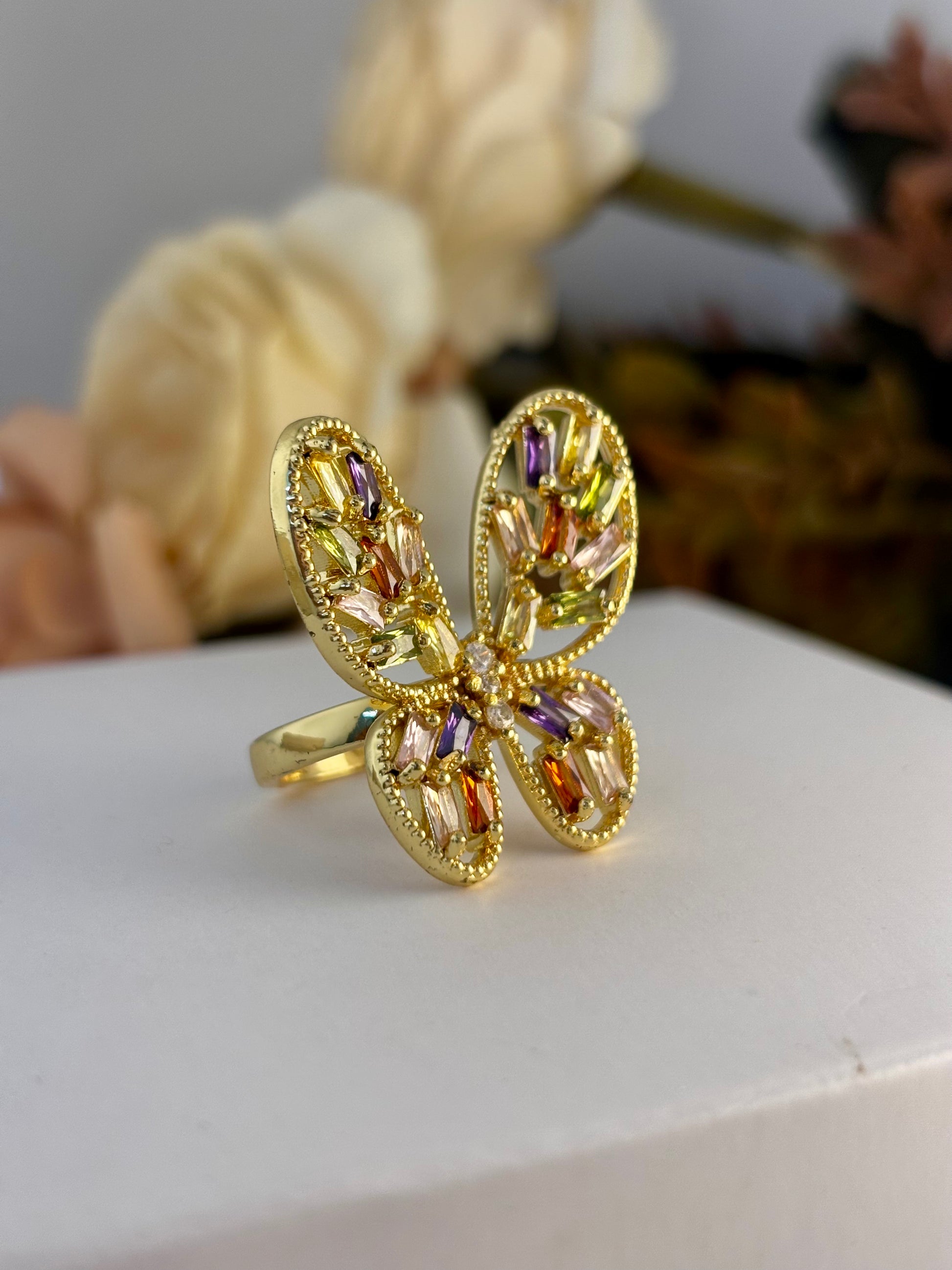 Gold butterfly ring with colorful gemstones on a white surface with flowers in the background