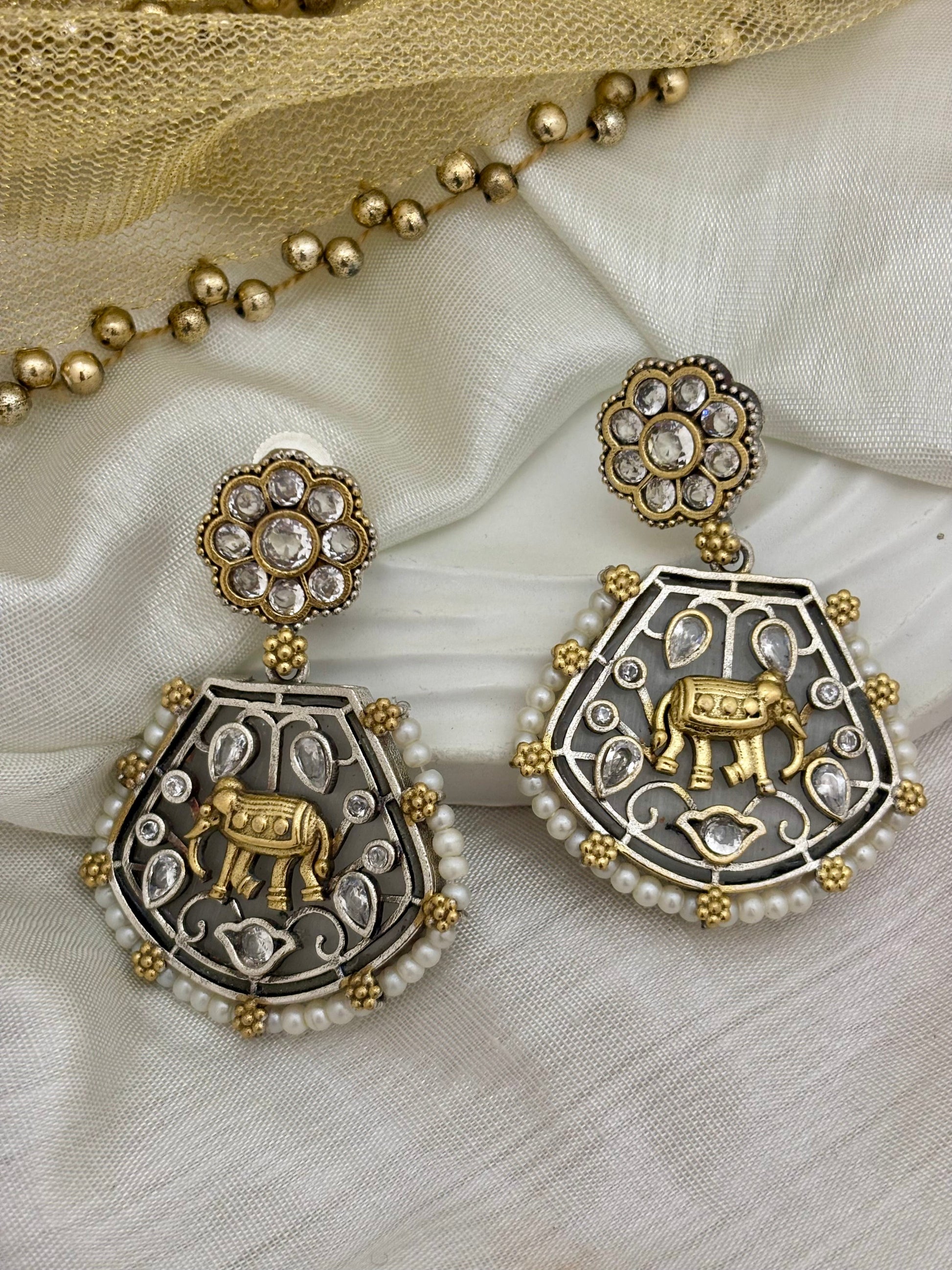 A pair of silver earrings with gold elephant motifs and white pearl fringes, displayed on a white fabric surface.