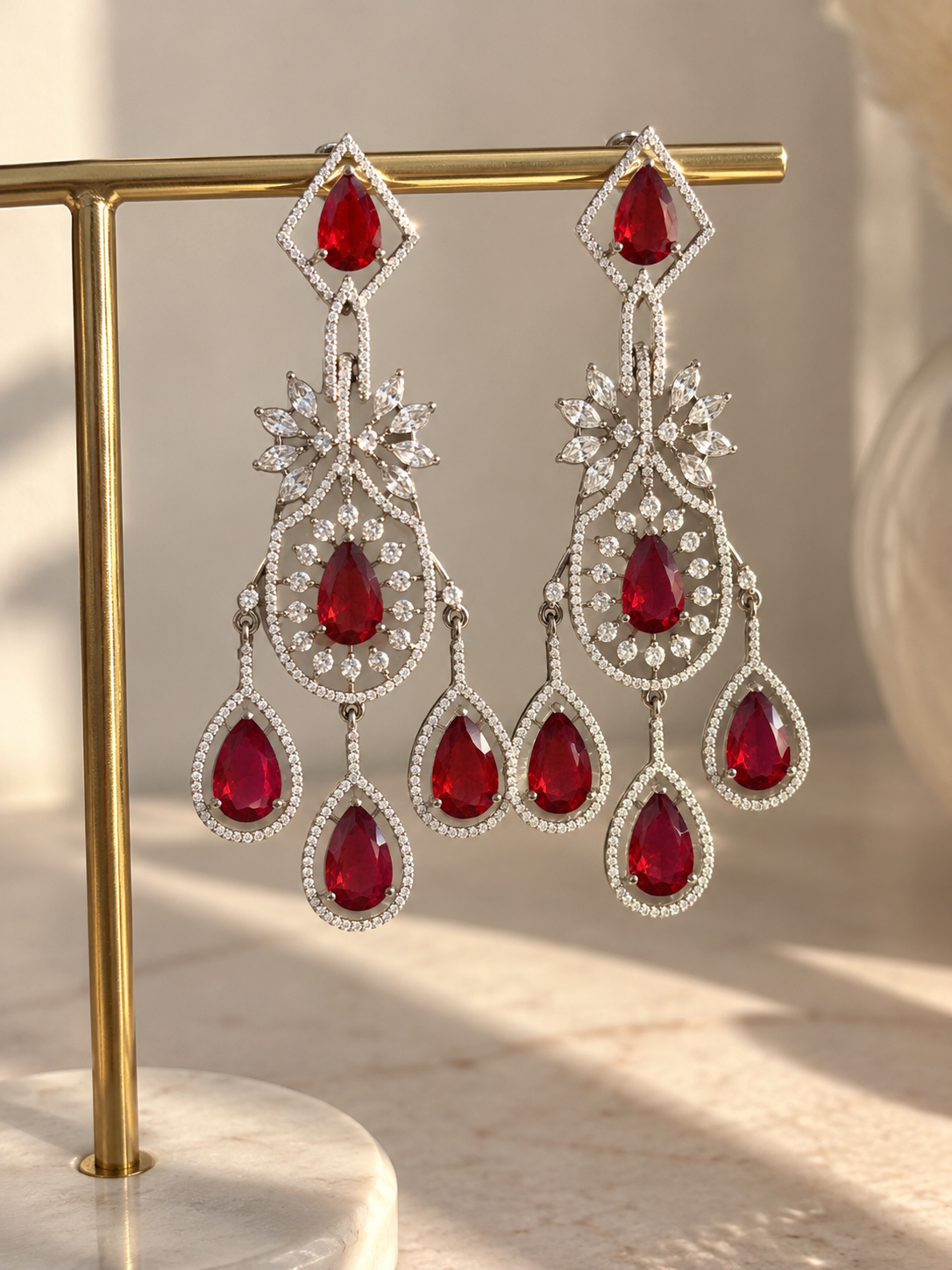 A pair of ornate silver-tone dangle earrings with large red teardrop gemstones and clear pavé accents, displayed on a gold stand against a softly lit neutral background.